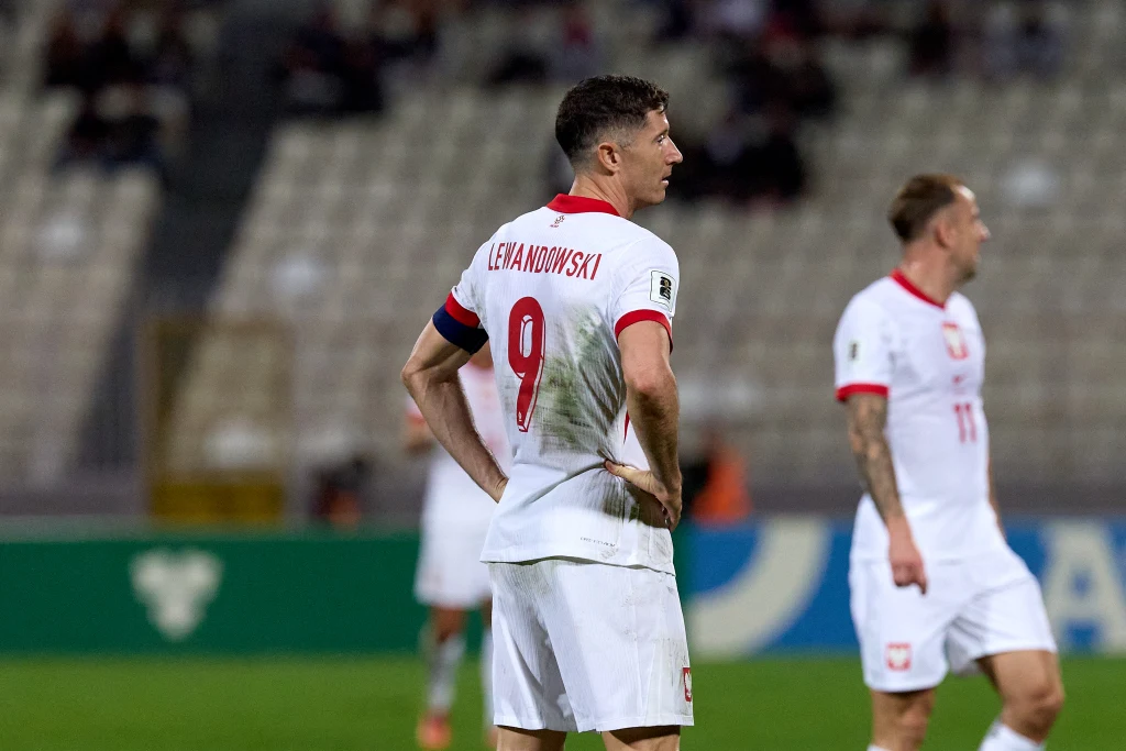 Piłkarz reprezentacji Polski z numerem 9 i nazwiskiem Lewandowski stoi na murawie podczas meczu, z widocznym śladem po kontakcie na koszulce, w tle drugi piłkarz również w stroju reprezentacyjnym.