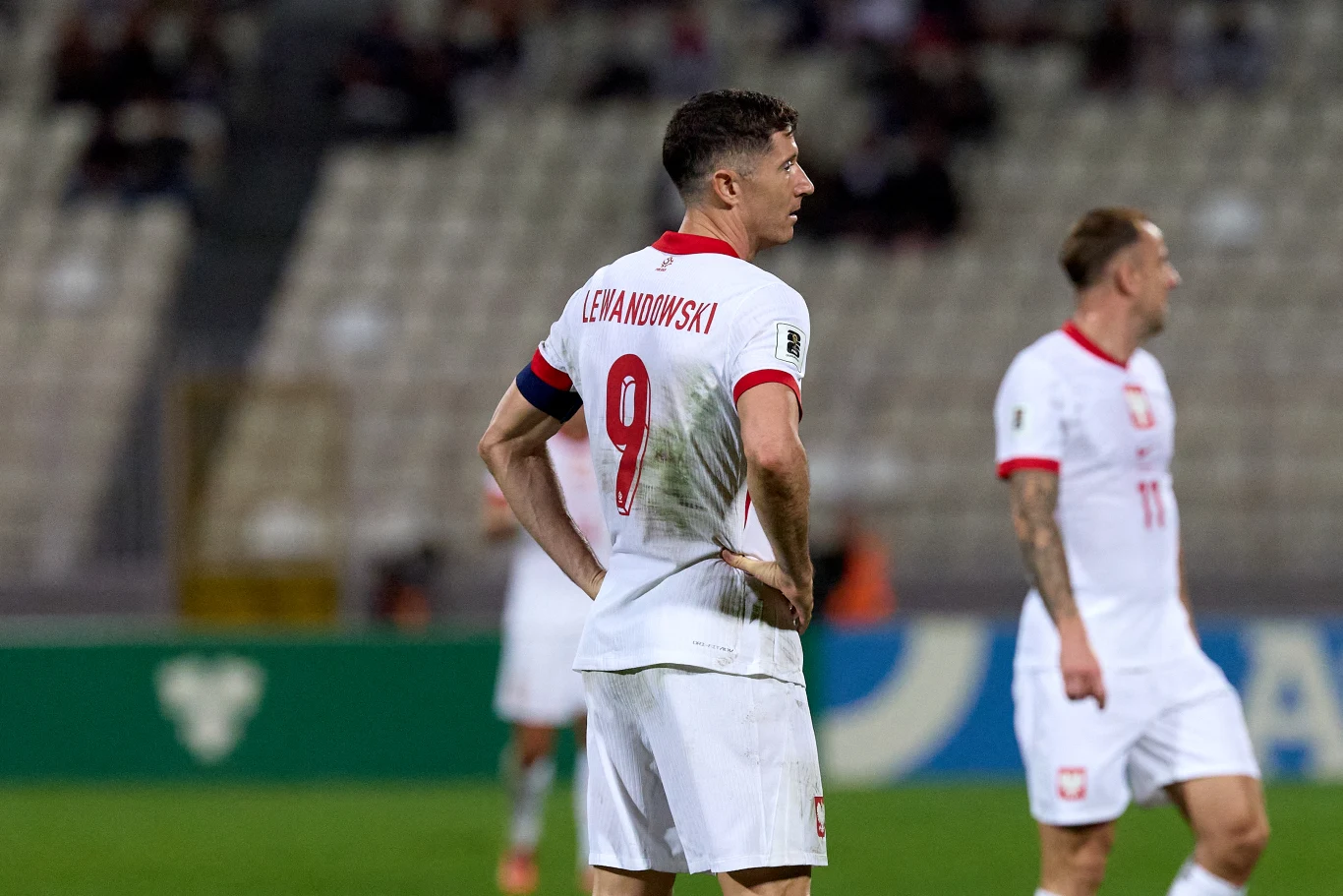 Piłkarz reprezentacji Polski z numerem 9 i nazwiskiem Lewandowski stoi na murawie podczas meczu, z widocznym śladem po kontakcie na koszulce, w tle drugi piłkarz również w stroju reprezentacyjnym.
