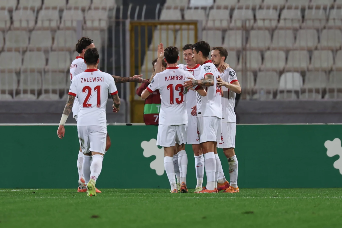 Grupa polskich piłkarzy świętuje zdobycie gola na stadionie, wszyscy ubrani w biało-czerwone stroje reprezentacji narodowej, wokół pustki na trybunach