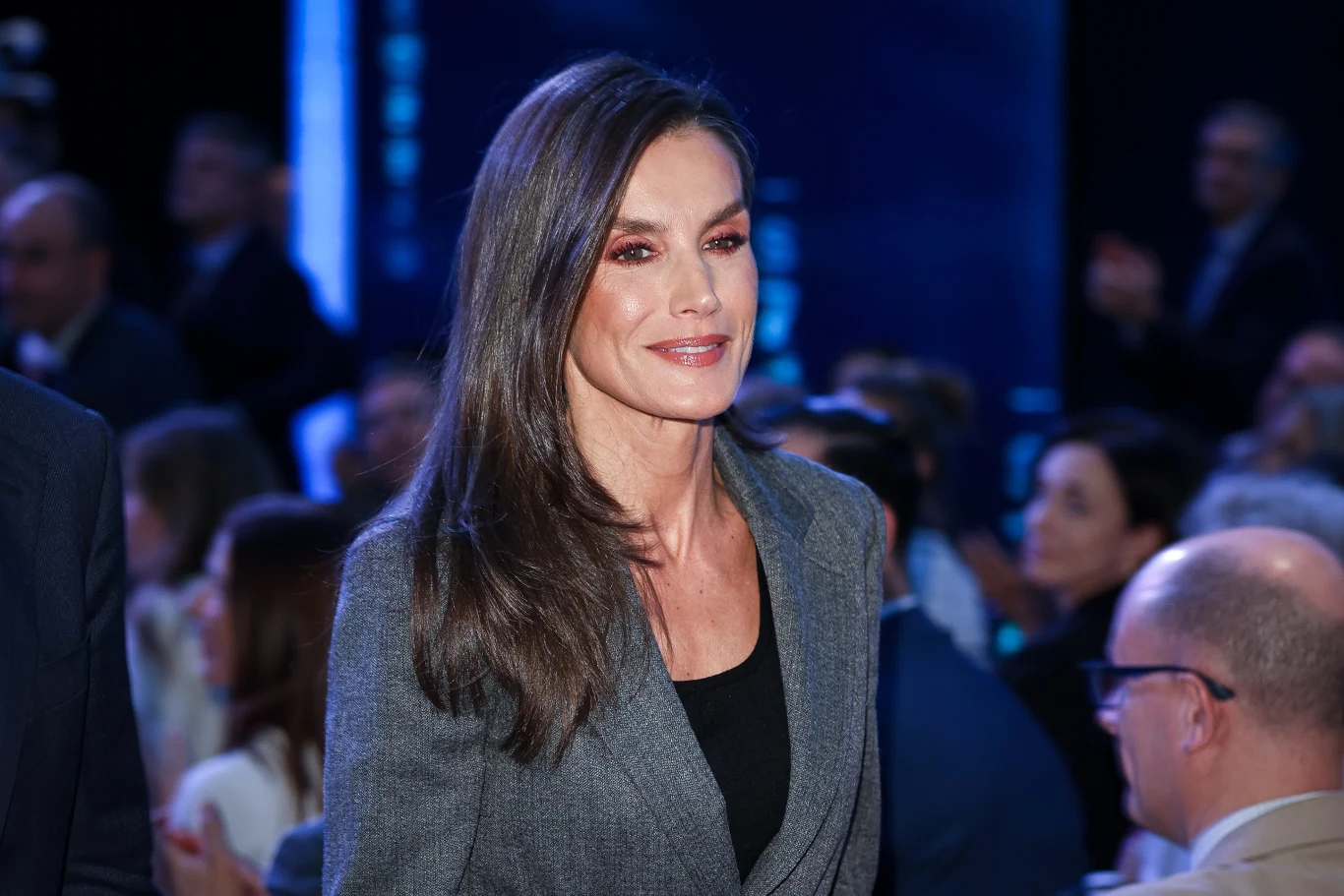 Królowa Letizia zachwyciła biurową stylizacją Elegancko ubrana kobieta z długimi, ciemnymi włosami stoi w centrum uwagi wśród grupy ludzi, jasno oświetlona na tle ciemniejszego, rozmytego tłumu.