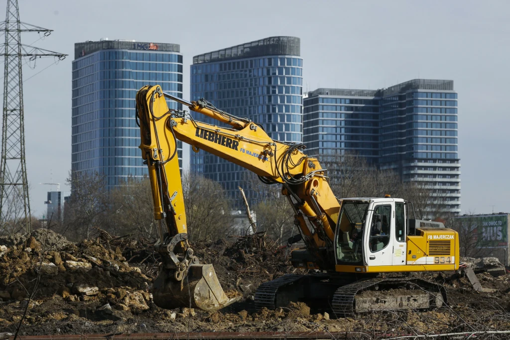 Firmy walczą z bankami o marże kredytowe Żółta koparka marki Liebherr pracująca na terenie budowy, wokół widoczne są ziemia i gruz, w tle nowoczesne szklane biurowce oraz linia energetyczna.