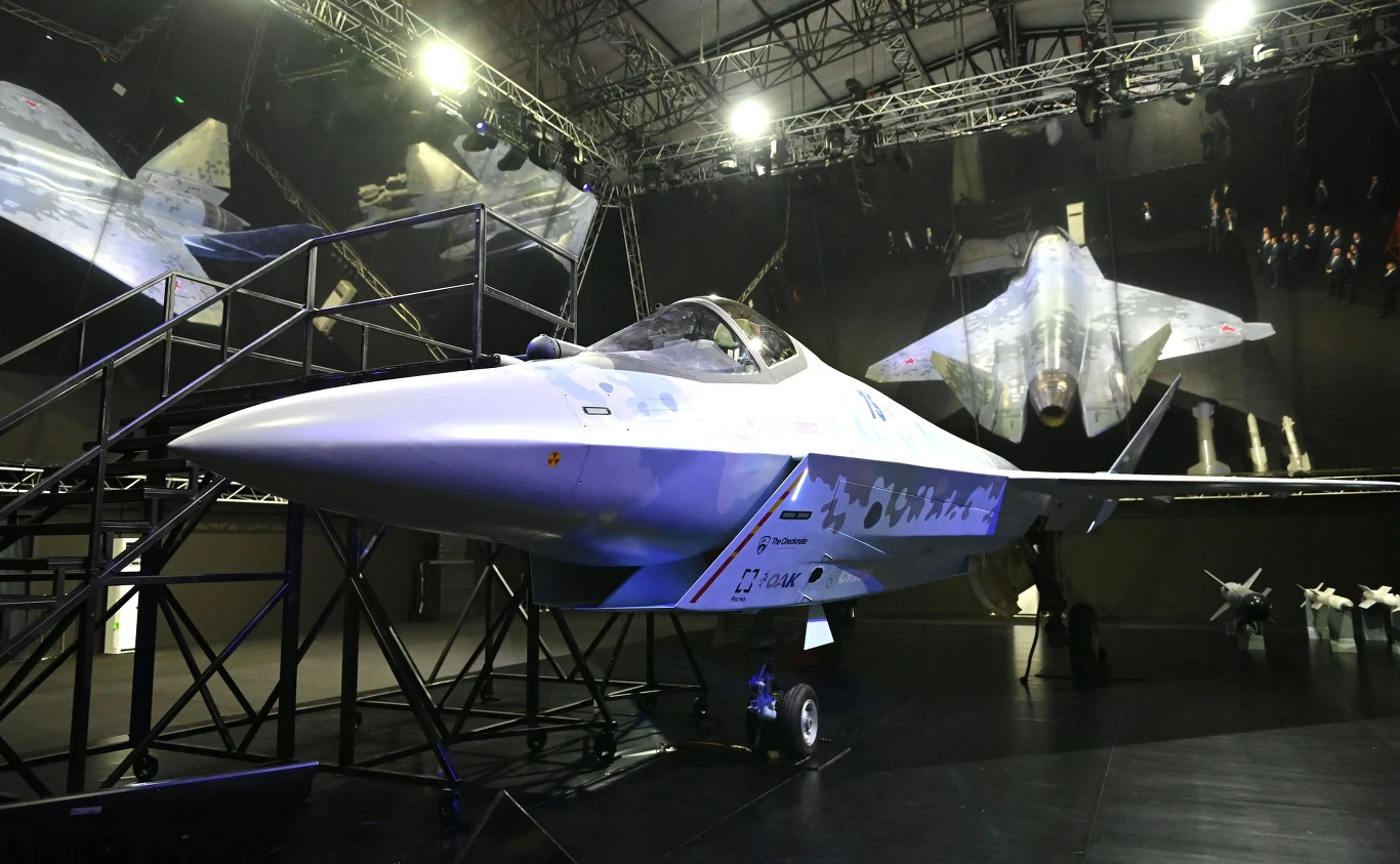 Model myśliwca Su-75 zaprezentowany na Międzynarodowym Salonie Lotniczo-Kosmicznym 2021 Nowoczesny myśliwiec odrzutowy prezentowany w hali wystawowej, oświetlony reflektorami, otoczony metalowymi barierkami. W tle widoczne są odbicia i grafiki innych samolotów na ścianach.