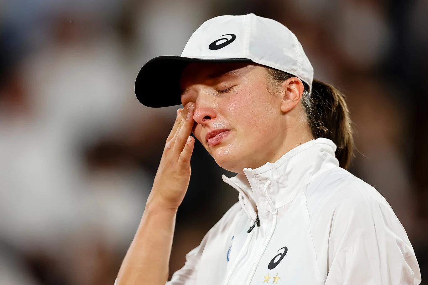 Iga Świątek Zawodniczka ubrana w sportowy strój i białą czapkę ociera łzę z oka, z wyrazem wzruszenia na twarzy, tło rozmyte, skupienie na emocjach osoby.