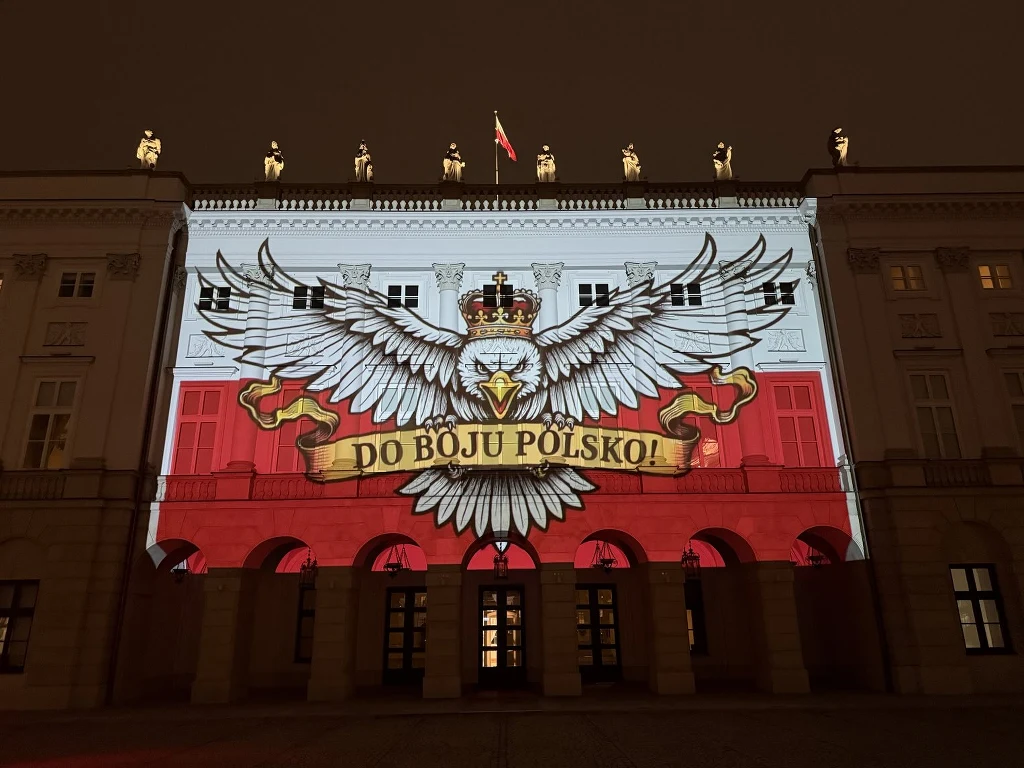 Fasada budynku oświetlona projekcją wizerunku białego orła z koroną, rozpostartymi skrzydłami oraz złotą wstęgą z napisem Do boju Polsko!, w tle widoczne barwy narodowe. Na dachu stoi osiem podświetlonych rzeźb oraz powiewa flaga Polski.