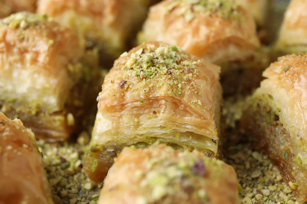 Baklava - co to takiego i jak przygotować w domu? Kawałki baklawy posypane zmielonymi pistacjami, ułożone blisko siebie na płaskiej powierzchni. Ciasto jest wielowarstwowe i mocno nasączone syropem, dzięki czemu błyszczy. Wierzch każdej porcji pokrywa chrupiąca, rumiana warstwa.