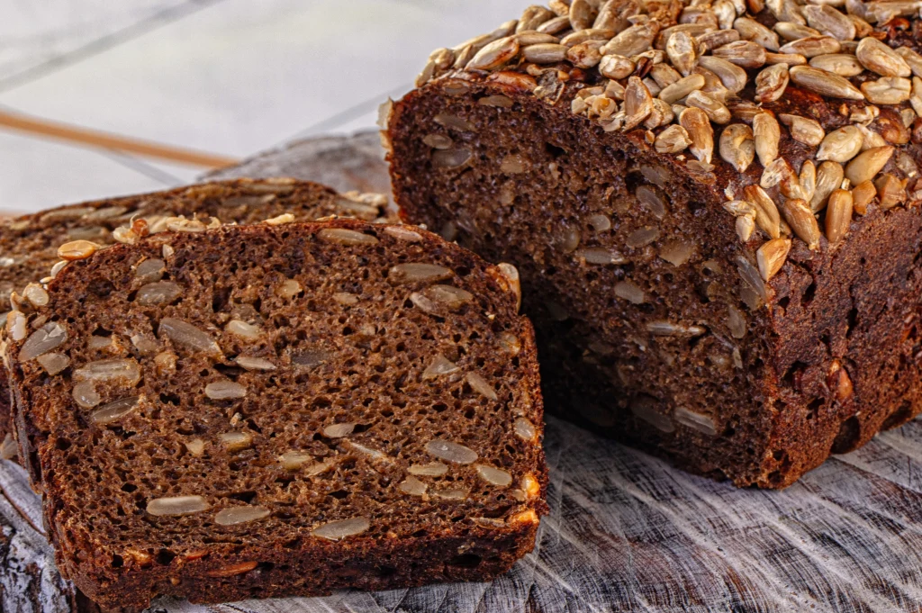 Pokrojony chleb razowy z ziarnami słonecznika, ciemne i gęste pieczywo, w przekroju widoczne liczne nasiona, wierzch bochenka posypany dużą ilością słonecznika, całość na drewnianej desce.