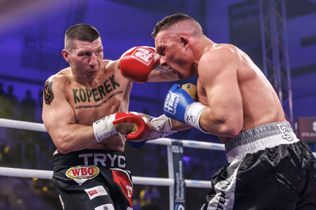 Mateusz Tryc (z lewej) Dwóch bokserów w ringu podczas dynamicznej wymiany ciosów, jeden z zawodników zadaje silny cios w twarz przeciwnika, napięte mięśnie i wyraz emocji podkreślają intensywność walki.