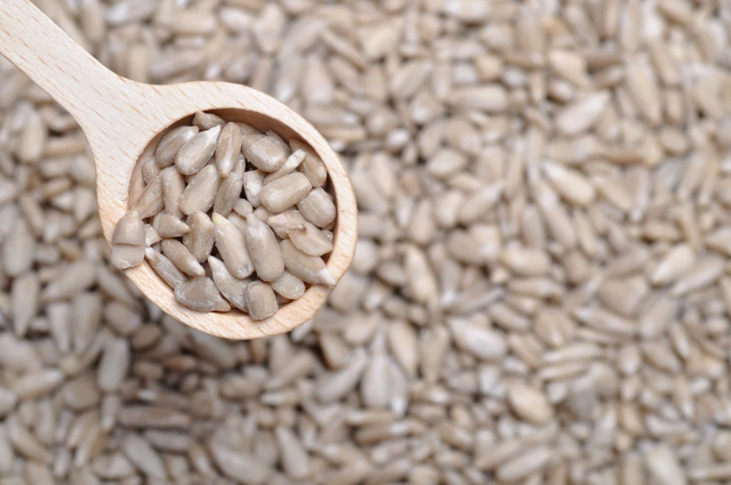 Drewniana łyżka wypełniona łuskanymi ziarnami słonecznika, leżąca na tle rozrzuconych ziaren słonecznika.