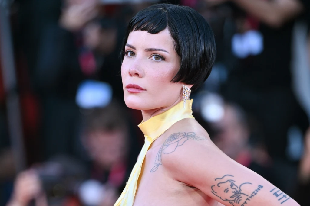 Halsey wystąpi na Open'er Festivalu Kobieta z krótkimi, czarnymi włosami ubrana w elegancką, żółtą suknię z odsłoniętymi ramionami. Ma widoczne tatuaże na ręce i ramieniu, nosi subtelne kolczyki, a jej twarz wyraża powagę. Rozmyte tło sugeruje obecność innych osób i atmosferę medialnego ...