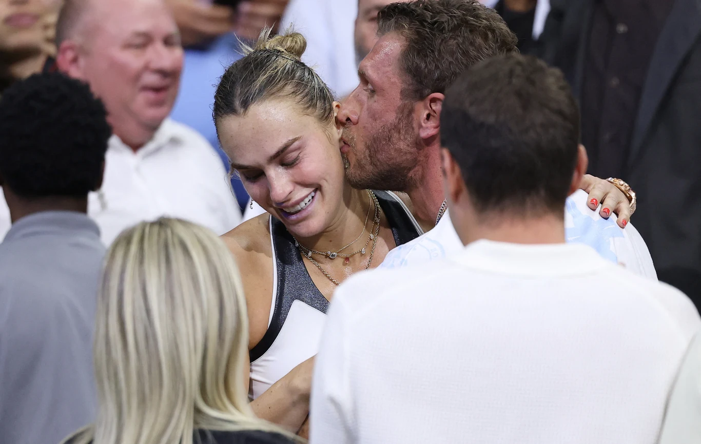 Sportowczyni ze wzruszeniem na twarzy przytulana jest przez mężczyznę, który całuje ją w policzek, wokół stoją inne osoby obserwujące tę intymną i radosną chwilę.