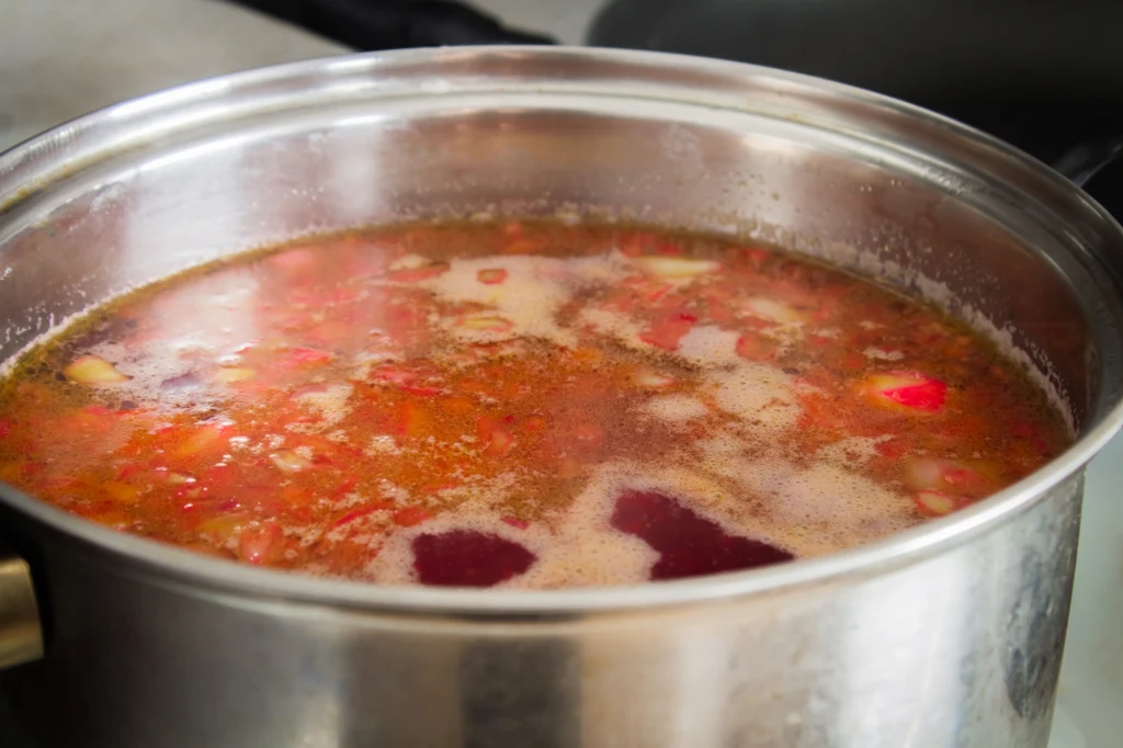 Garnek z gotującym się barszczem z widocznymi kawałkami buraków oraz warzyw, na powierzchni lekka piana.