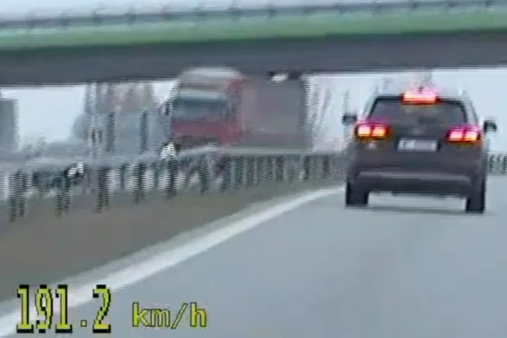 Czarny samochód jedzie z dużą prędkością autostradą, na liczniku pomiaru widoczna prędkość 191,2 km/h. Na jezdni wyraźne oznakowanie pasa ruchu oraz w tle ciężarówka i wiadukt.