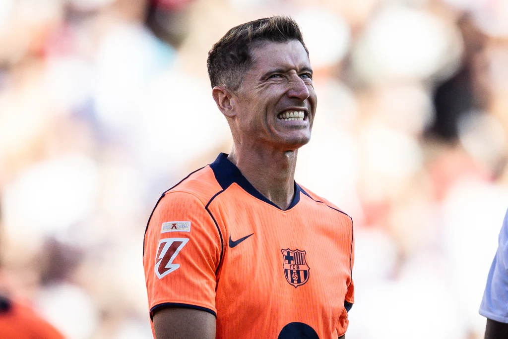Robert Lewandowski Piłkarz ubrany w pomarańczową koszulkę z herbem FC Barcelony uśmiecha się szeroko, na tle rozmazanego stadionu i tłumu.