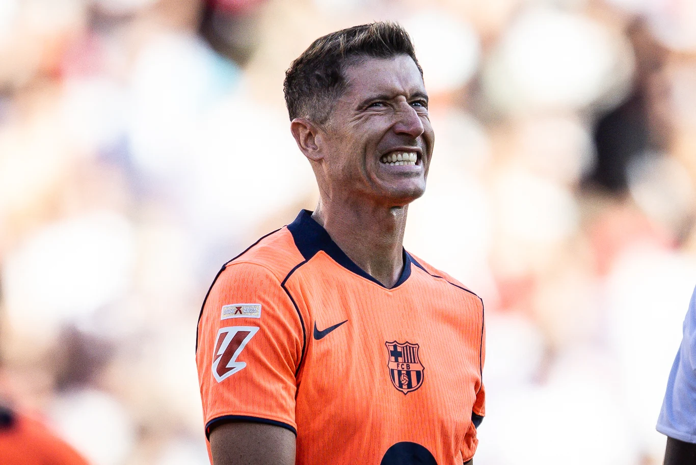 Piłkarz ubrany w pomarańczową koszulkę z herbem FC Barcelony uśmiecha się szeroko, na tle rozmazanego stadionu i tłumu.