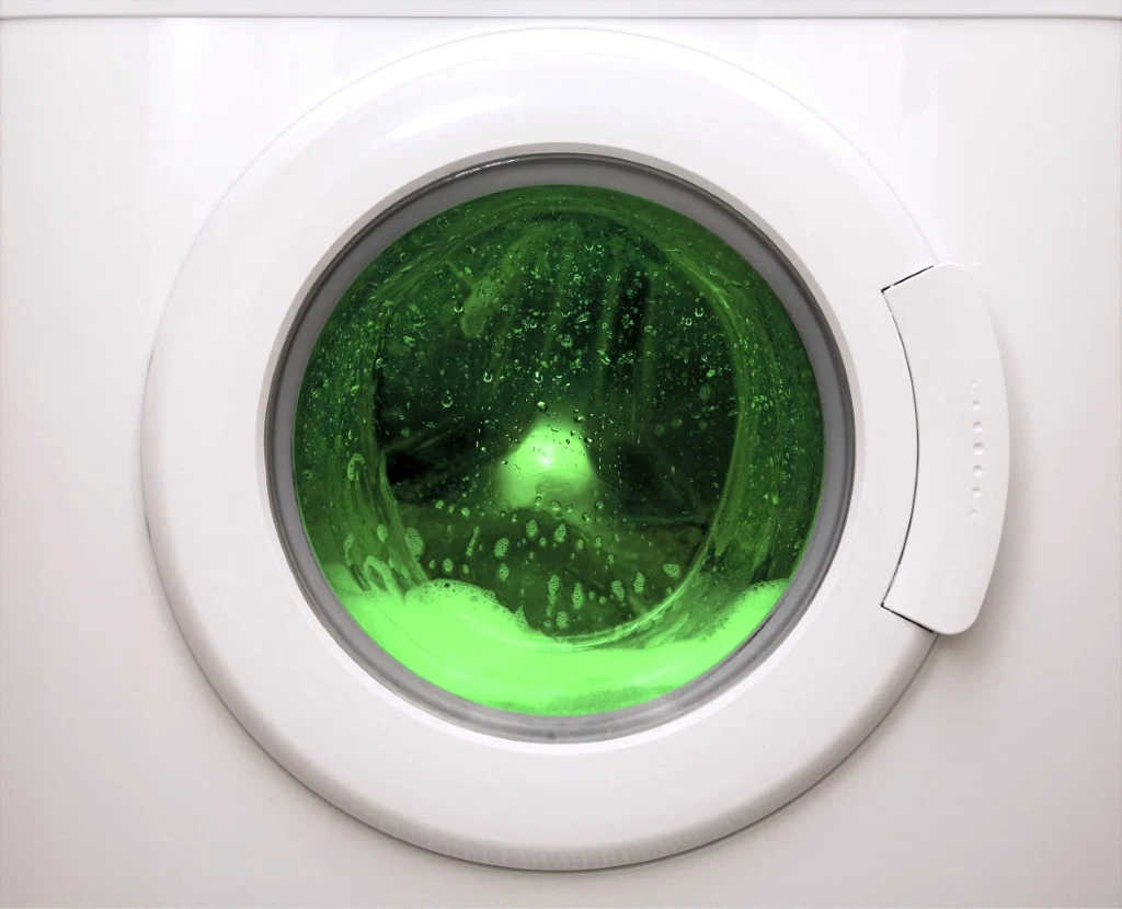 Bęben pralki z przeszklonymi drzwiczkami, w środku intensywnie zielona piana i woda, co nadaje nietypowy, kolorowy efekt.