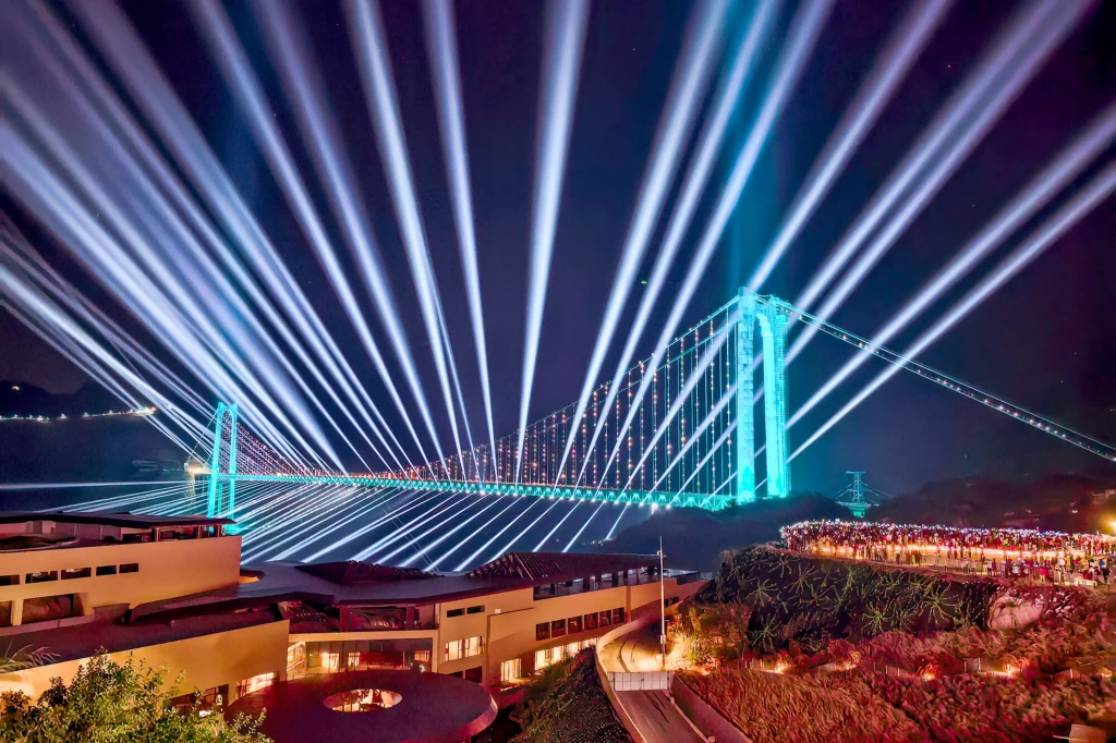 Huajiang Grand Canyon Bridge trafił do Księgi Rekordów Guinnessa Most podświetlony kolorowymi reflektorami i laserami, z których snopy światła rozchodzą się w niebo. W tle widoczne rozświetlone niebo nocne oraz duża grupa widzów obserwujących widowisko z pobliskiego tarasu.