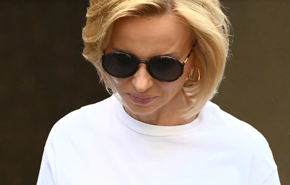 Kobieta z blond włosami w krótkiej fryzurze, ubrana w białą koszulkę i ciemne okulary przeciwsłoneczne, patrzy w dół. W tle ciemna, rozmyta powierzchnia.