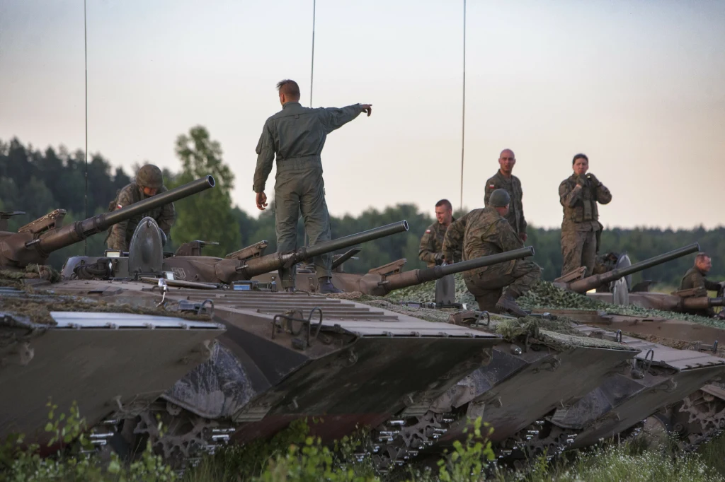 Polscy żołnierze podczas ćwiczeń w Orzyszu, zdj. ilustracyjne Grupa żołnierzy w mundurach stoi oraz siedzi na czołgach ustawionych w rzędzie na tle leśnego krajobrazu, jeden z nich wskazuje ręką kierunek, pozostali rozmawiają lub obserwują otoczenie.