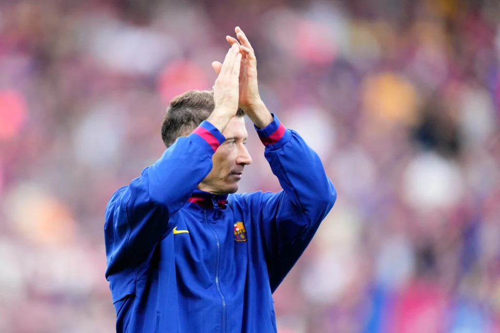 Robert Lewandowski Mężczyzna w niebieskiej sportowej kurtce z herbem FC Barcelony klaszcze, zwrócony bokiem do kamery, w tle rozmyte trybuny stadionu.