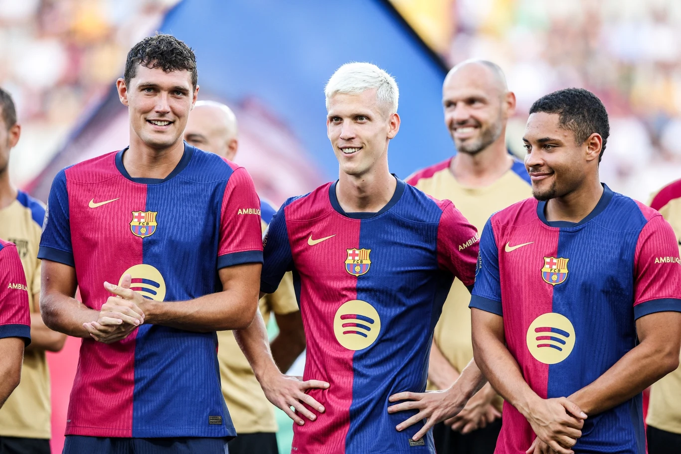 Trzech piłkarzy FC Barcelony w oficjalnych strojach drużyny stoi obok siebie na murawie, uśmiechają się i patrzą w jednym kierunku, w tle znajdują się inni członkowie drużyny.