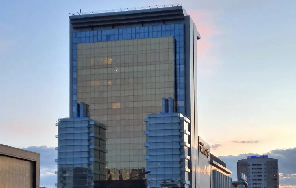 Nowoczesny biurowiec z dużą, złotą fasadą odbijającą niebo, otoczony elementami architektonicznymi z przeszklonymi bokami. Na tle widać inne wysokie budynki oraz wieczorne niebo z delikatnym różowym światłem.