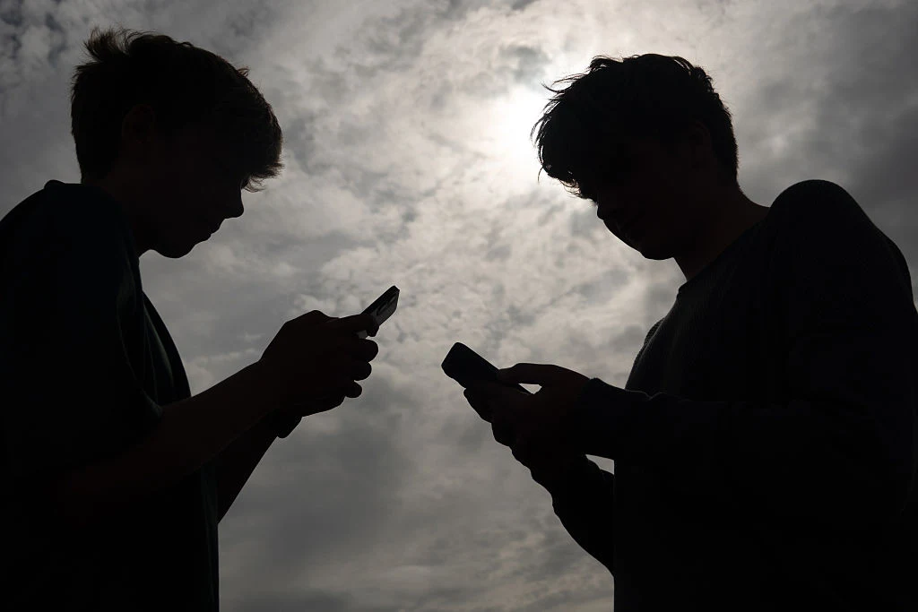 Dzieci spędzające czas przed ekranami telefonów Dwie sylwetki osób stojących naprzeciwko siebie z telefonami w dłoniach, oświetlone od tyłu przez pochmurne niebo z widocznym słońcem.