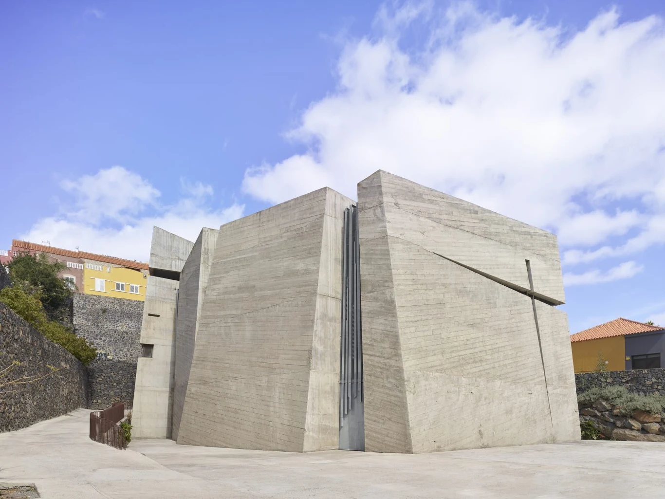 Betonowy kościół na Teneryfie został najlepszym budynkiem świata Nowoczesny budynek z betonu o surowej, geometrycznej formie, ustawiony na otwartym placu, otoczony niższą zabudową mieszkalną oraz murkami z kamieni. W tle błękitne niebo z lekko rozproszonymi chmurami.