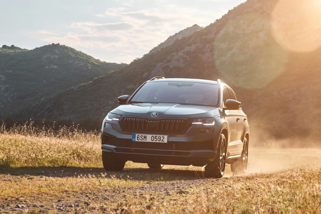 Napęd 4x4 wygląda na gadżet dla miłośników wymagających tras, ale w praktyce staje się wsparciem również w zwyczajnych sytuacjach Czarny SUV marki Škoda jedzie polną drogą wśród wysokiej trawy, na tle gór oświetlonych promieniami zachodzącego słońca.
