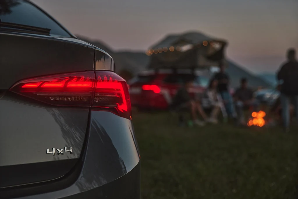 Napęd 4x4 to nie tylko zabawka dla fanów ekstremalnych tras Tył samochodu terenowego z oznaczeniem 4x4 na tle rozświetlonych tylnych świateł, w dalszym planie drugi pojazd z namiotem dachowym oraz grupa osób siedzących wokół ogniska, wszystko w otoczeniu przyrody i w wieczornej atmosferze.