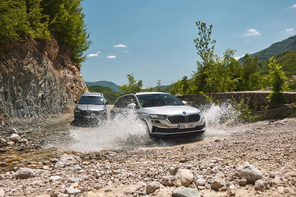 Napęd 4x4 to wsparcie, które sprawdza się w każdych warunkach – od miejskich ulic po leśne ścieżki Dwa samochody terenowe jadą przez płytki strumień, rozpryskując wodę, otoczone przez kamienisty szlak i górski krajobraz z zielonymi drzewami i niebem bez chmur.