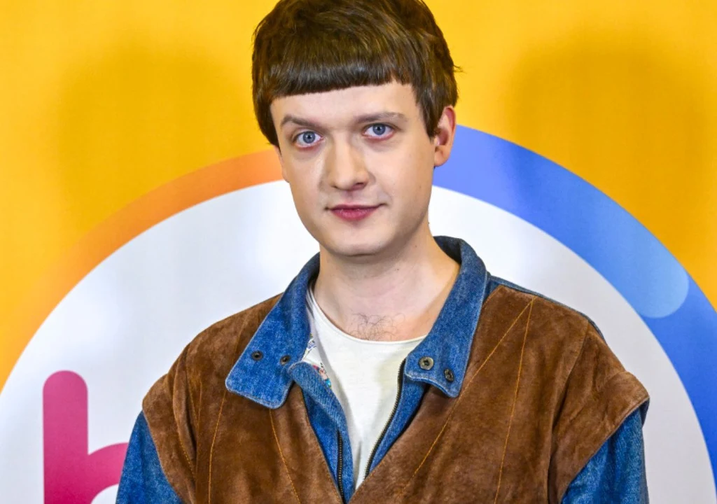 Młody mężczyzna o jasnej cerze z krótką fryzurą, ubraną w brązowo-niebieską kurtkę z dżinsu i zamszu, stoi na żółtym tle z kolorowym logotypem.