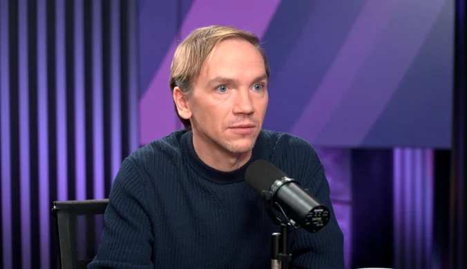 Mężczyzna w jasnych włosach siedzący przy stole, ubrany w granatowy sweter, mówiący do profesjonalnego mikrofonu na tle fioletowych i czarnych paneli wygłuszających.