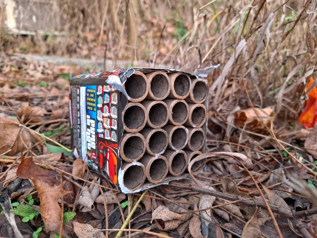 Puste opakowanie po fajerwerkach leży na ziemi wśród suchych liści i gałęzi.