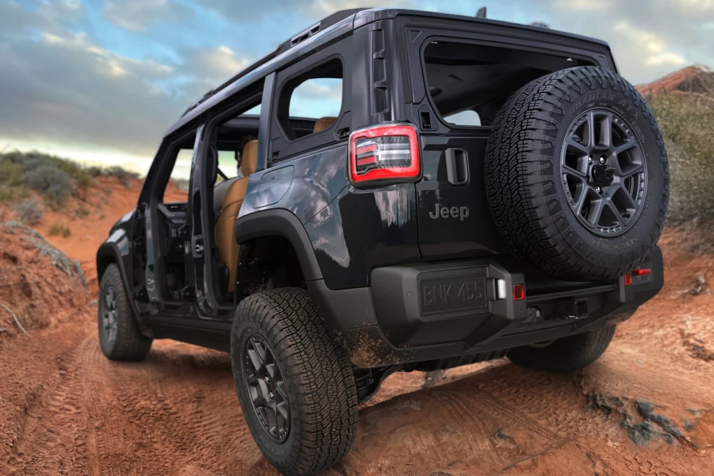 Czarny samochód terenowy Jeep z otwartymi drzwiami jedzie po piaszczystej, wyboistej drodze terenowej wśród niskiej roślinności. Na dachu widoczne relingi, a na tylnej klapie zamocowane jest koło zapasowe.