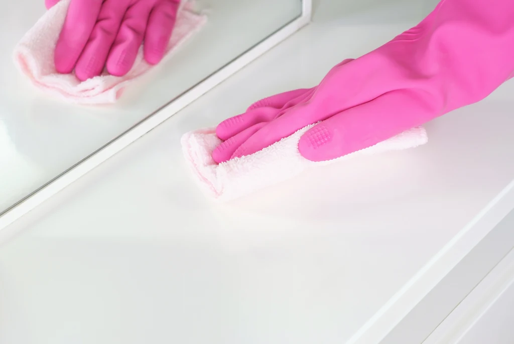 Różowa rękawiczka na dłoni trzymającej białą ścierkę, czyszczącej jasną powierzchnię przed lustrem.