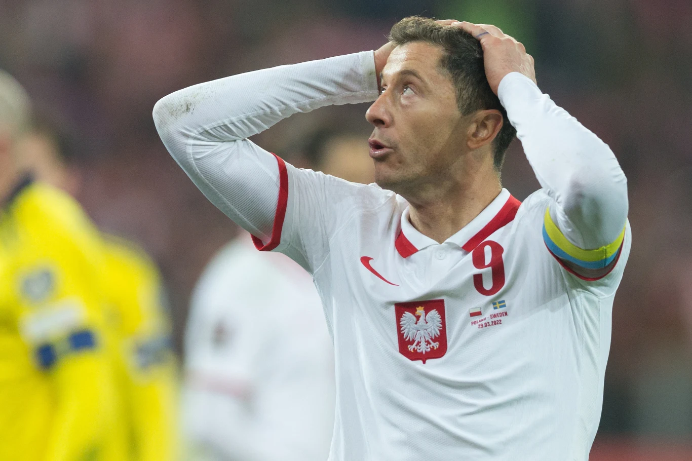 Robert Lewandowski Piłkarz w białej koszulce z orłem i numerem 9 oraz opaską kapitana trzyma ręce na głowie i wyraża zaskoczenie lub rozczarowanie podczas meczu, w tle rozmyte postacie sędziów i zawodników.