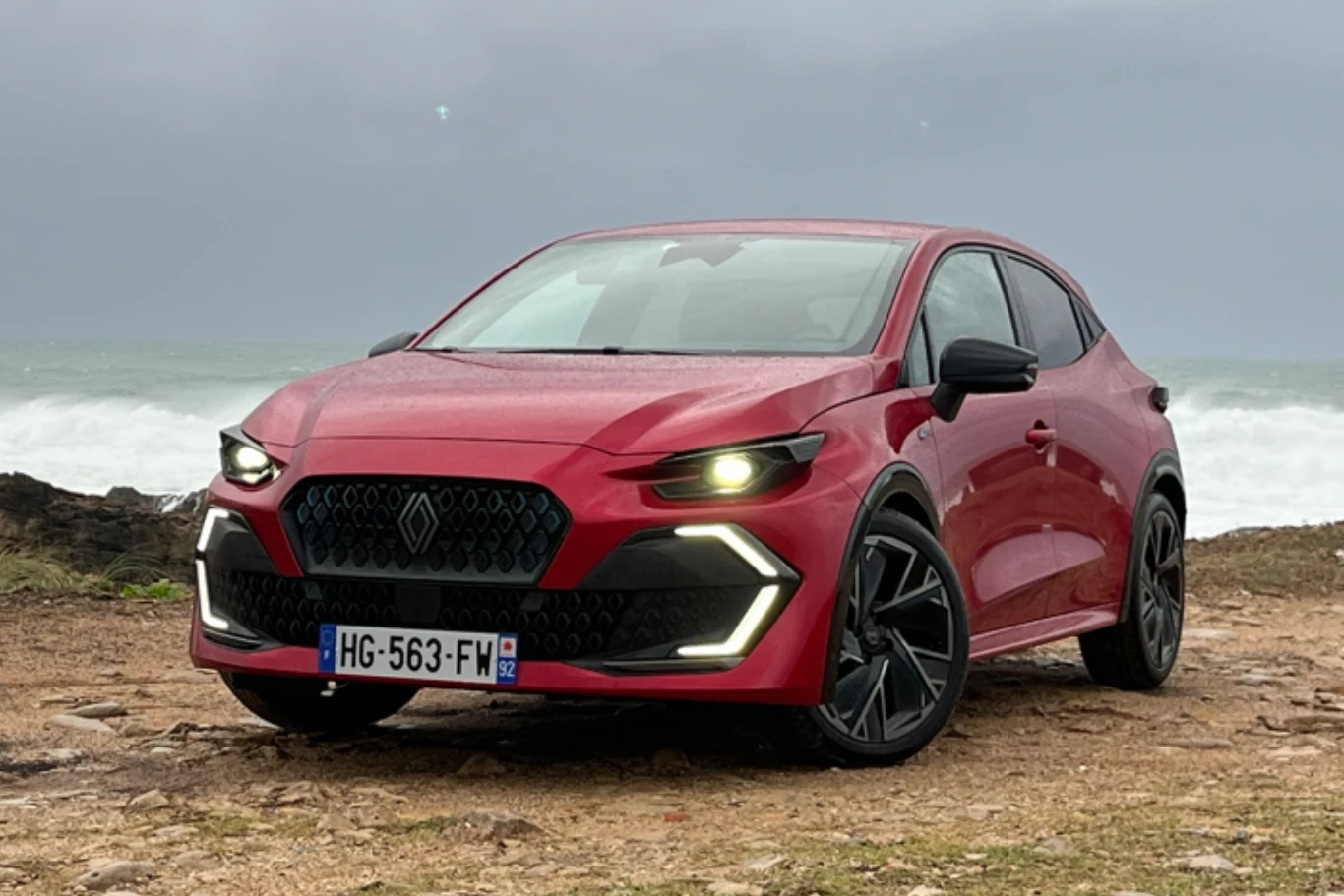 Czerwony samochód osobowy o nowoczesnym wyglądzie stoi na skraju wybrzeża, na jasnym, ziemistym podłożu, z włączonymi światłami do jazdy dziennej w kształcie litery L. W tle wzburzone morze i pochmurne niebo.