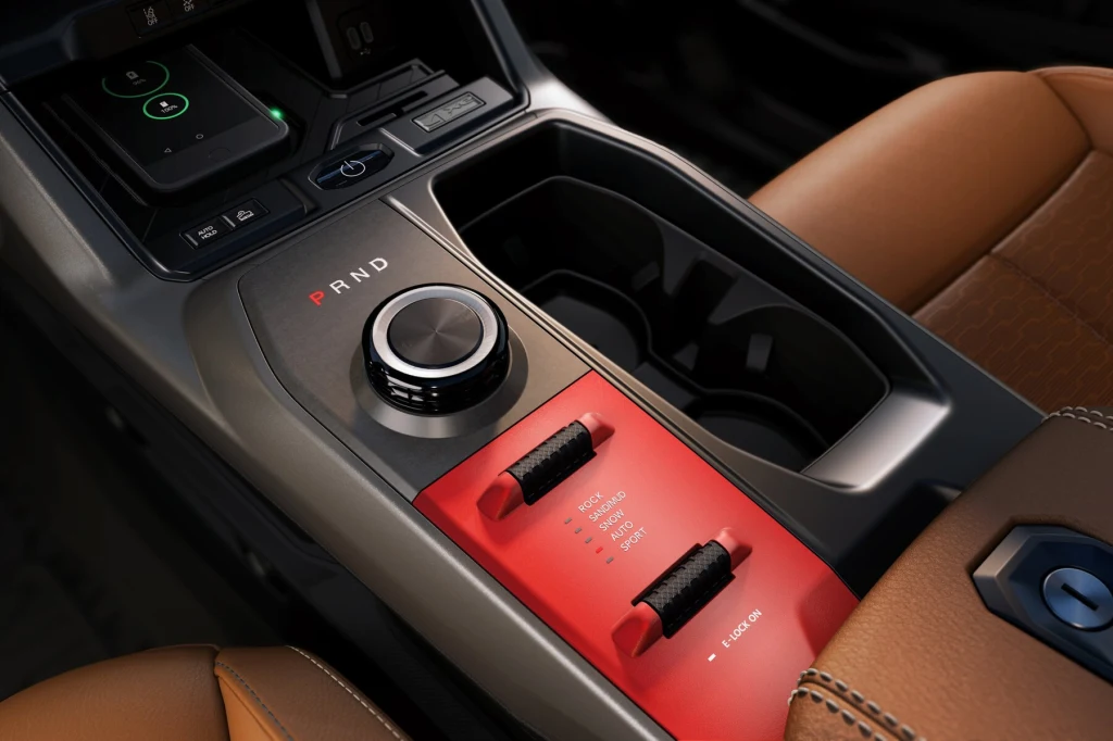 Nowoczesna konsola środkowa samochodu z pokrętłem zmiany biegów, dwoma czarnymi suwakami, uchwytami na napoje i fragmentem brązowego fotela wykonanego ze skóry.