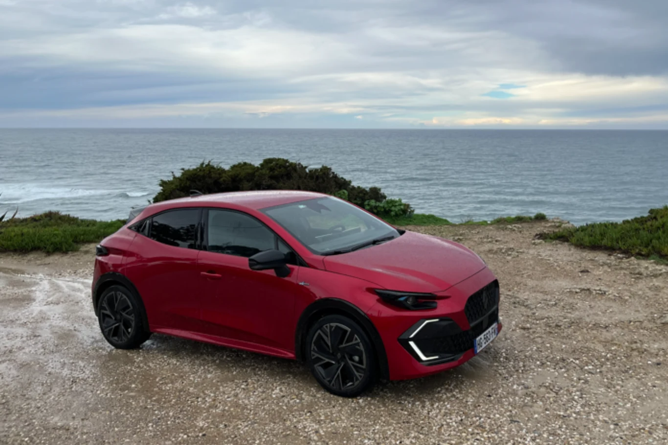 Nowoczesny czerwony samochód osobowy ustawiony na żwirowym podłożu z widokiem na morze, w tle niskie krzewy i pochmurne niebo.