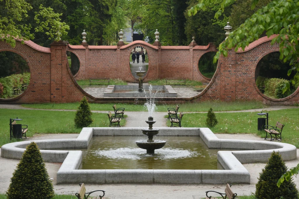 Zadbana fontanna o geometrycznym kształcie otoczona zieloną trawą i symetrycznie ustawionymi ławkami, z tyłu charakterystyczna ceglasta ściana z okrągłymi otworami prowadząca do kolejnej części ogrodu.