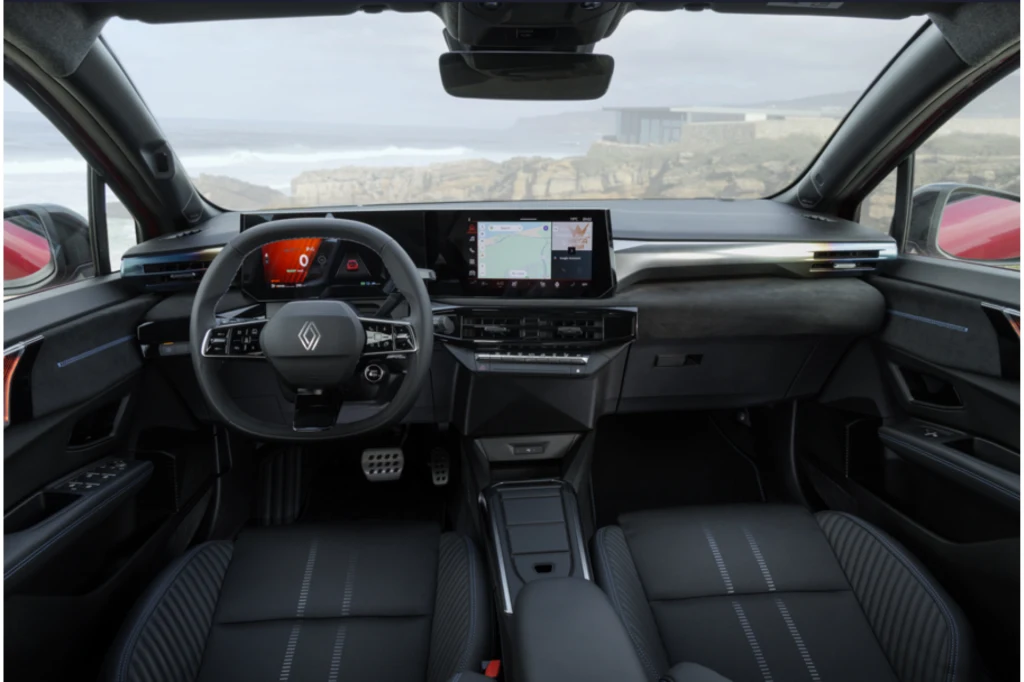 Wnętrze nowoczesnego samochodu marki Renault z widokiem na kokpit, kierownicę, dwa duże wyświetlacze cyfrowe oraz wygodne fotele pokryte czarną tapicerką. Przez szyby widoczny fragment wybrzeża oraz rozmyte tło krajobrazu.