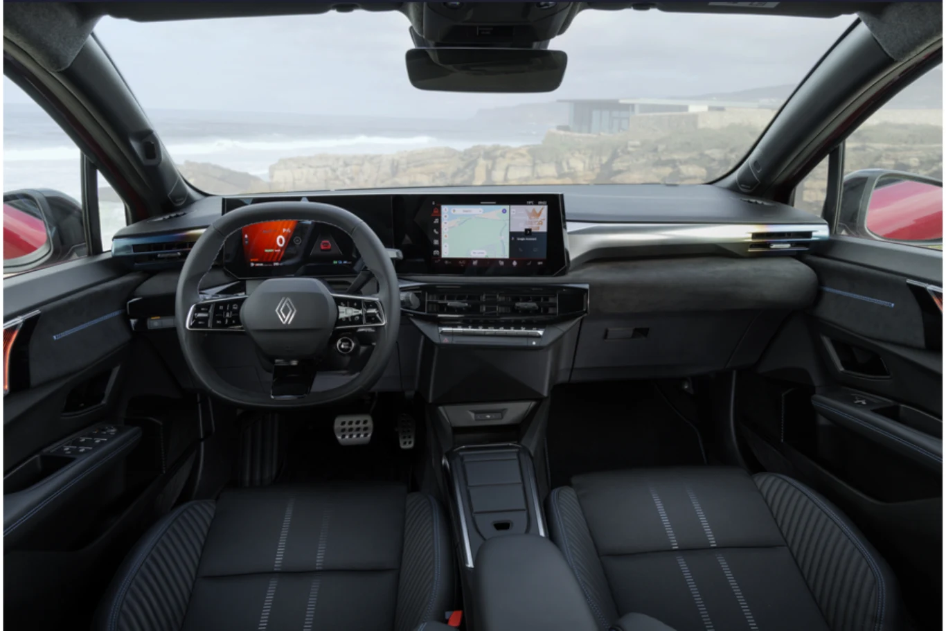 Wnętrze nowoczesnego samochodu marki Renault z widokiem na kokpit, kierownicę, dwa duże wyświetlacze cyfrowe oraz wygodne fotele pokryte czarną tapicerką. Przez szyby widoczny fragment wybrzeża oraz rozmyte tło krajobrazu.