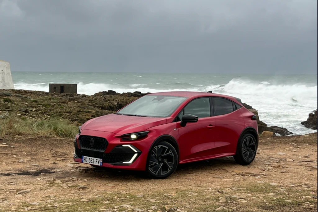 Czerwony samochód osobowy sportowego typu SUV zaparkowany na wybrzeżu przy wzburzonym morzu, z widocznymi nowoczesnymi reflektorami LED i ciemnymi alufelgami, w tle szare, pochmurne niebo oraz fale rozbijające się o skały.