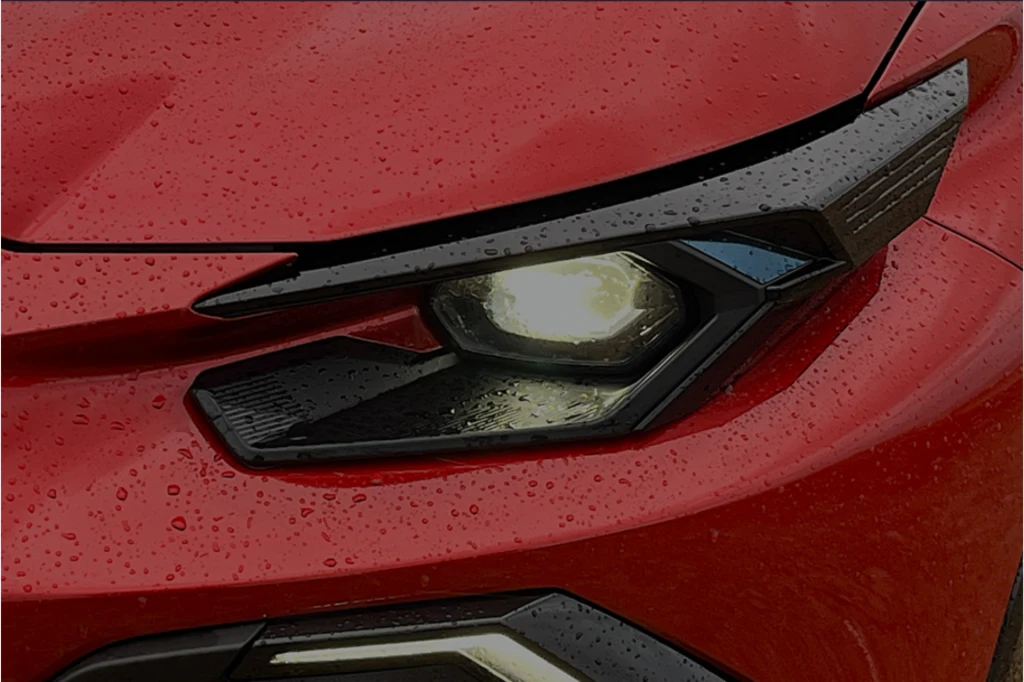 Detail czerwonego samochodu z widocznym przednim reflektorem włączonym, na powierzchni karoserii widać krople deszczu.