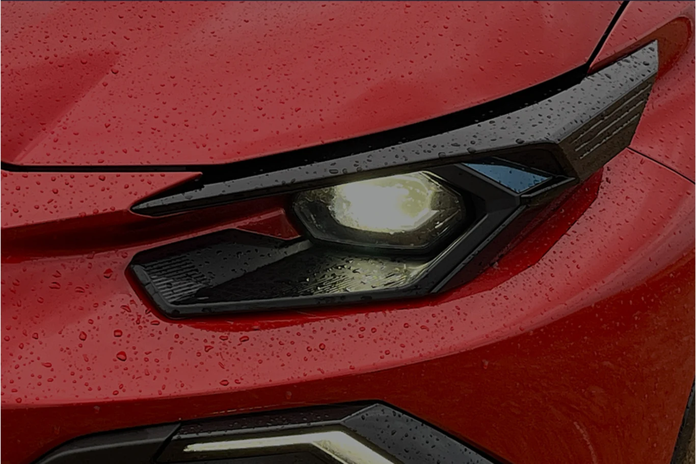Detail czerwonego samochodu z widocznym przednim reflektorem włączonym, na powierzchni karoserii widać krople deszczu.