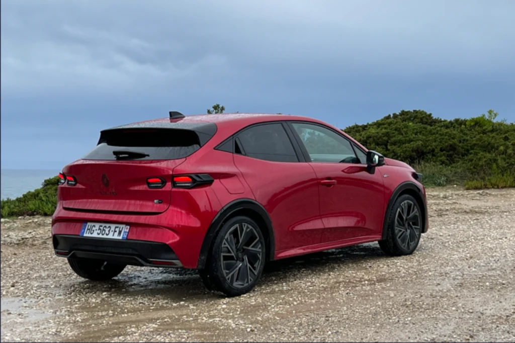 Czerwony samochód typu SUV stoi na żwirowej drodze przy zielonych zaroślach, na tle pochmurnego nieba. Auto skierowane tyłem, widoczne czarne felgi oraz francuska tablica rejestracyjna.