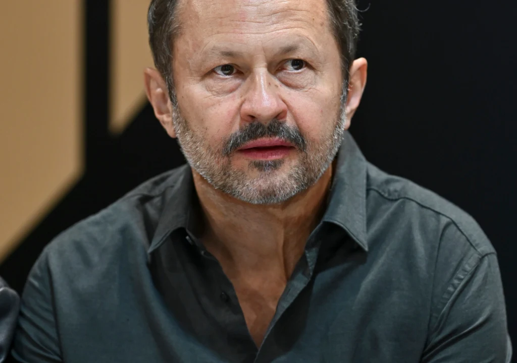 Mężczyzna w średnim wieku o krótkich, ciemnych włosach i brodzie, ubrany w ciemną koszulę, siedzi na tle czarnej i beżowej ściany. Jego twarz ukazuje skupienie lub zamyślenie.