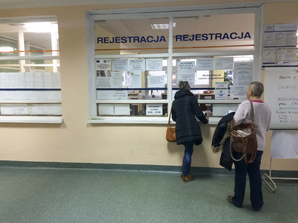 Dwa kobiety stoją przy okienku rejestracji w budynku, najprawdopodobniej w placówce medycznej. Ściana wokół okienka jest wypełniona informacjami i ulotkami, a wnętrze po drugiej stronie okna wygląda jak część biura.
