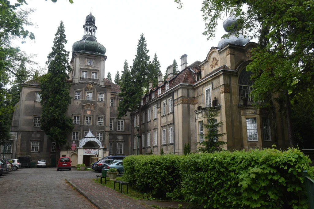 Historyczny budynek z wysoką wieżą i klasyczną architekturą otoczony drzewami, z zaparkowanymi samochodami na podjeździe oraz bujną zielenią wzdłuż alei wejściowej.