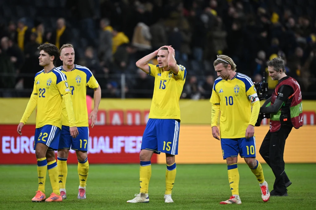 Reprezentacja Szwecji Czterech piłkarzy reprezentacji Szwecji w żółto-niebieskich strojach opuszcza boisko wyrażając rozczarowanie po meczu, jeden z nich trzyma głowę w geście frustracji, a w tle widoczny jest stadion z kibicami.